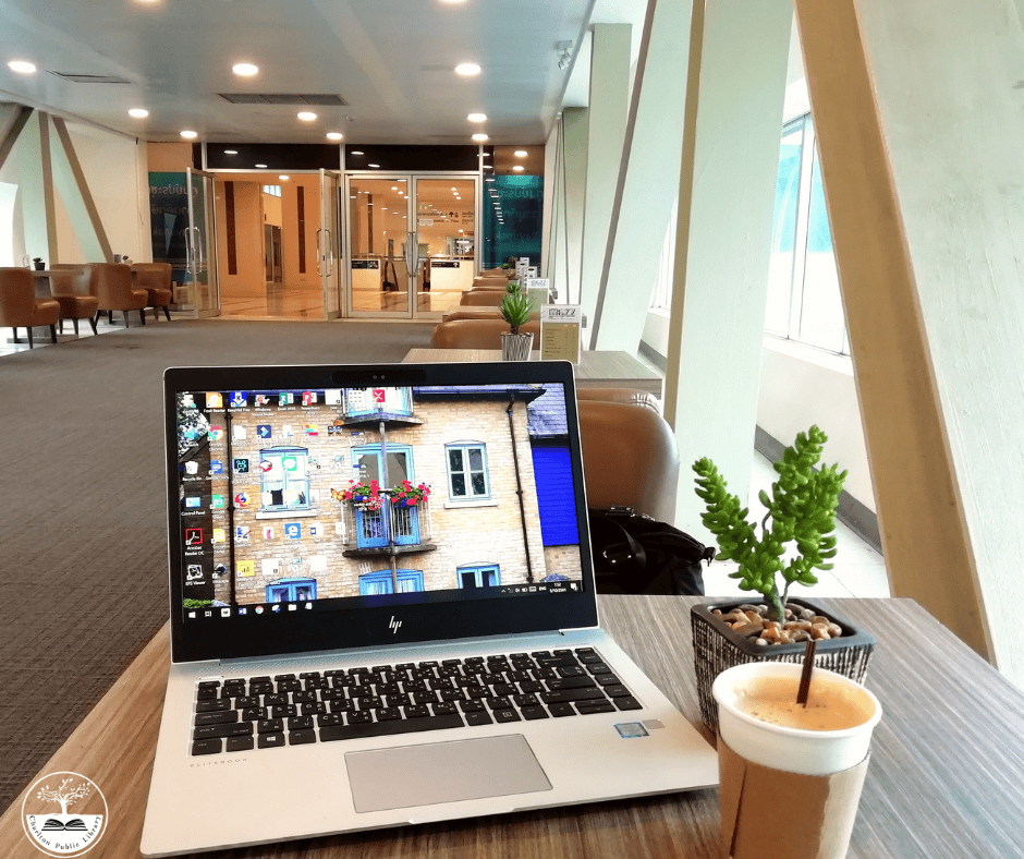 a laptop, iced coffee, and small potted plant on a table in a modern lounge area with large windows and seating in the background.