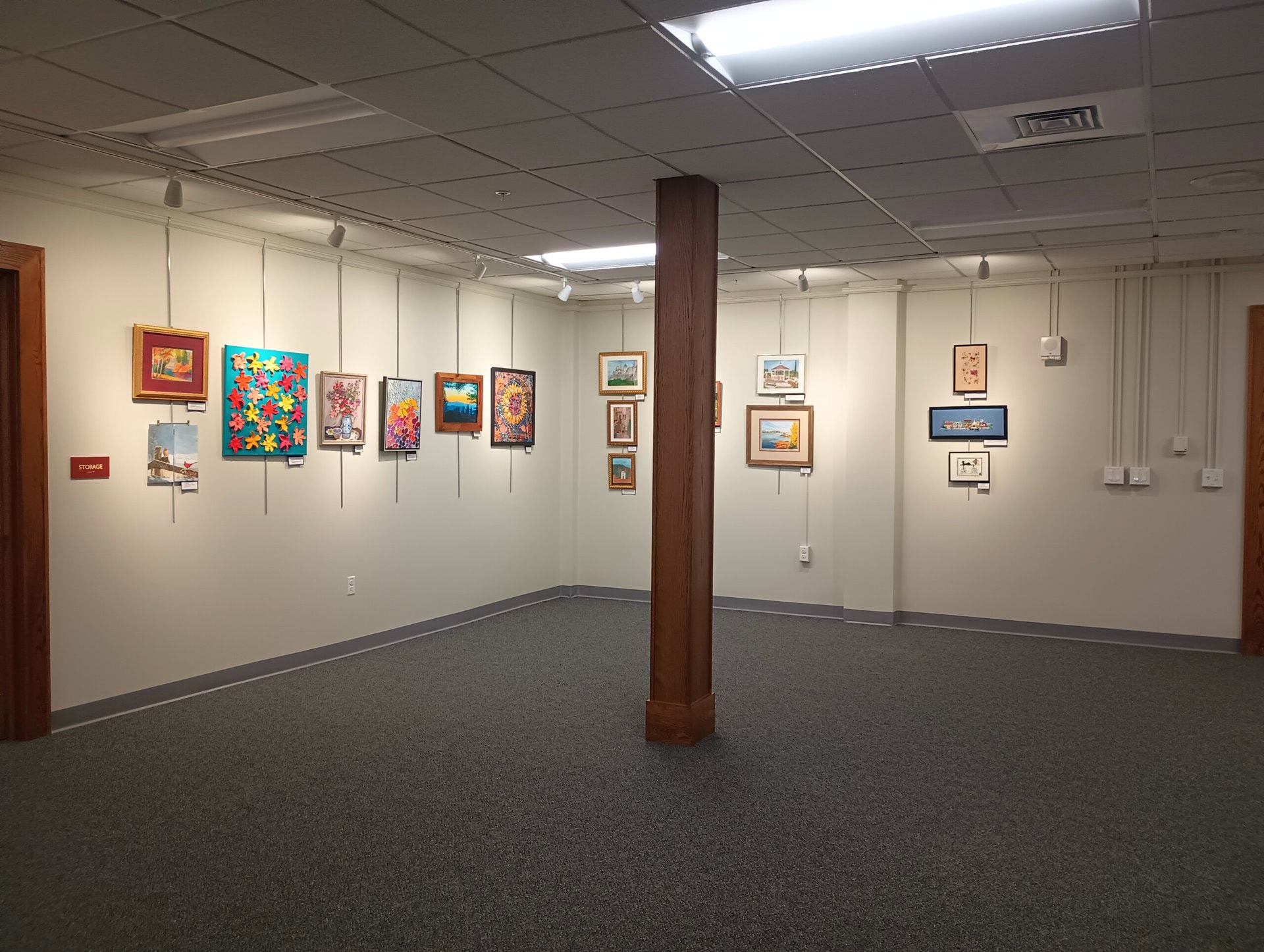 an art gallery room with various framed paintings displayed on cream colored walls, gray carpet flooring, a wooden support column, and overhead fluorescent lighting.