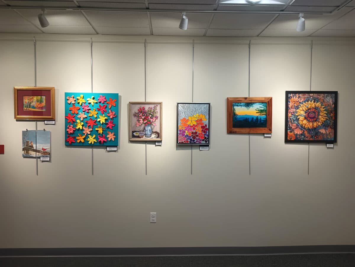 seven colorful paintings of various styles and subjects are displayed on a white gallery wall, each individually framed and lit from above by ceiling spotlights.