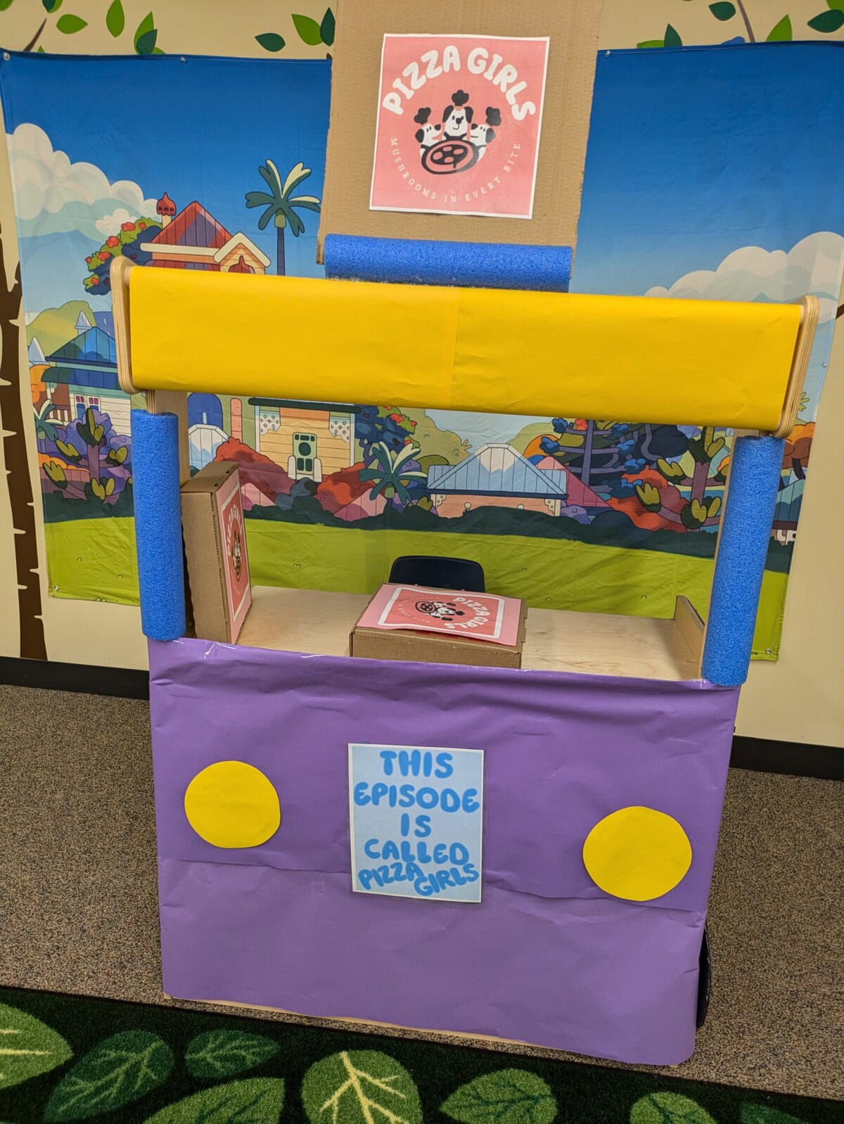 A colorful cardboard booth with a "Pizza Girls" sign, yellow and purple decorations, and a blue sign reading "This episode is called Pizza Girls.