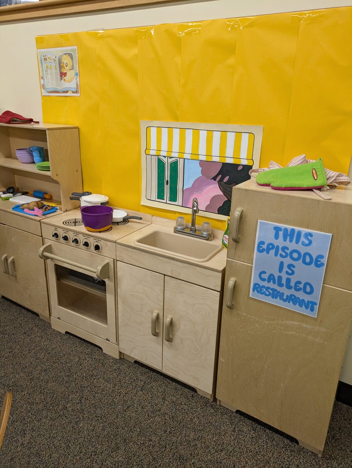 Preschool play kitchen setup with toy stove, sink, dishes, and a sign reading "THIS EPISODE IS CALLED RESTAURANT" on a yellow background.