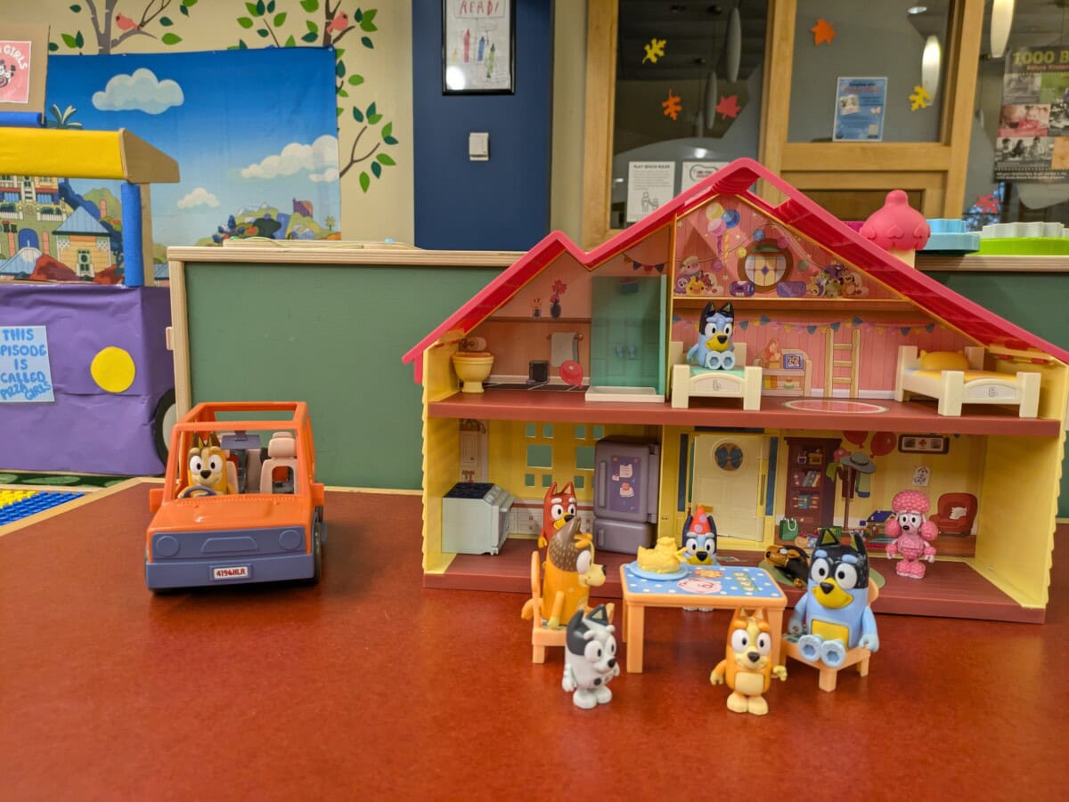 A Bluey dollhouse playset with figurines and a toy car is displayed on a red table in a colorful playroom setting.