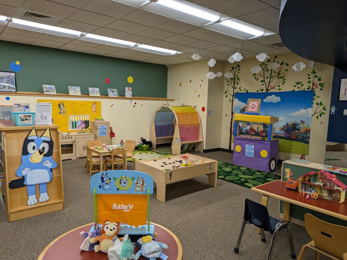 A colorful daycare room with tables, chairs, a playhouse, toys, wall decorations, and cartoon character displays, including Bluey near the entrance.