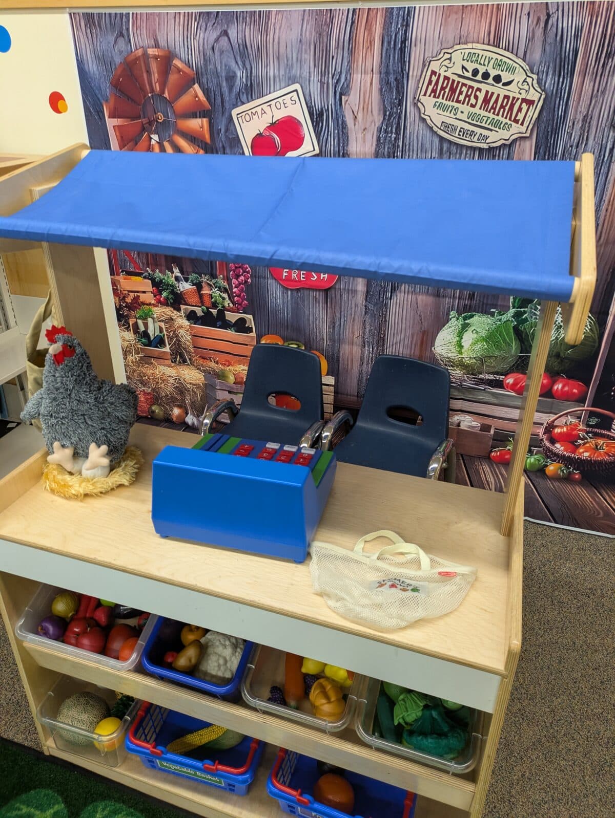 a children's play market setup with toy fruits, vegetables, a cash register, two chairs, and a plush chicken, in front of a farmers market themed backdrop.