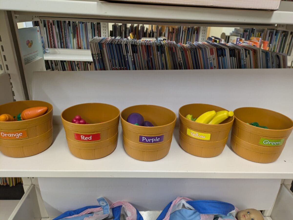 five brown baskets labeled orange, red, purple, yellow, and green contain matching colored toy fruits and vegetables, displayed on a shelf with books in the background.