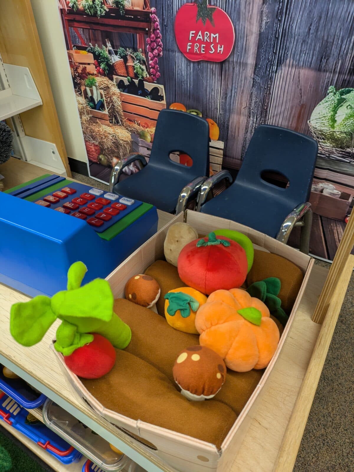 a play grocery store setup with plush vegetables in a wooden crate, a blue toy cash register, two child sized chairs, and a "farm fresh" sign in the background.